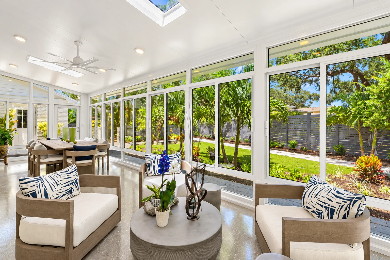A bright all-season room with floor-to-ceiling windows, a skylight, and neutral lounge seating overlooks a green garden.