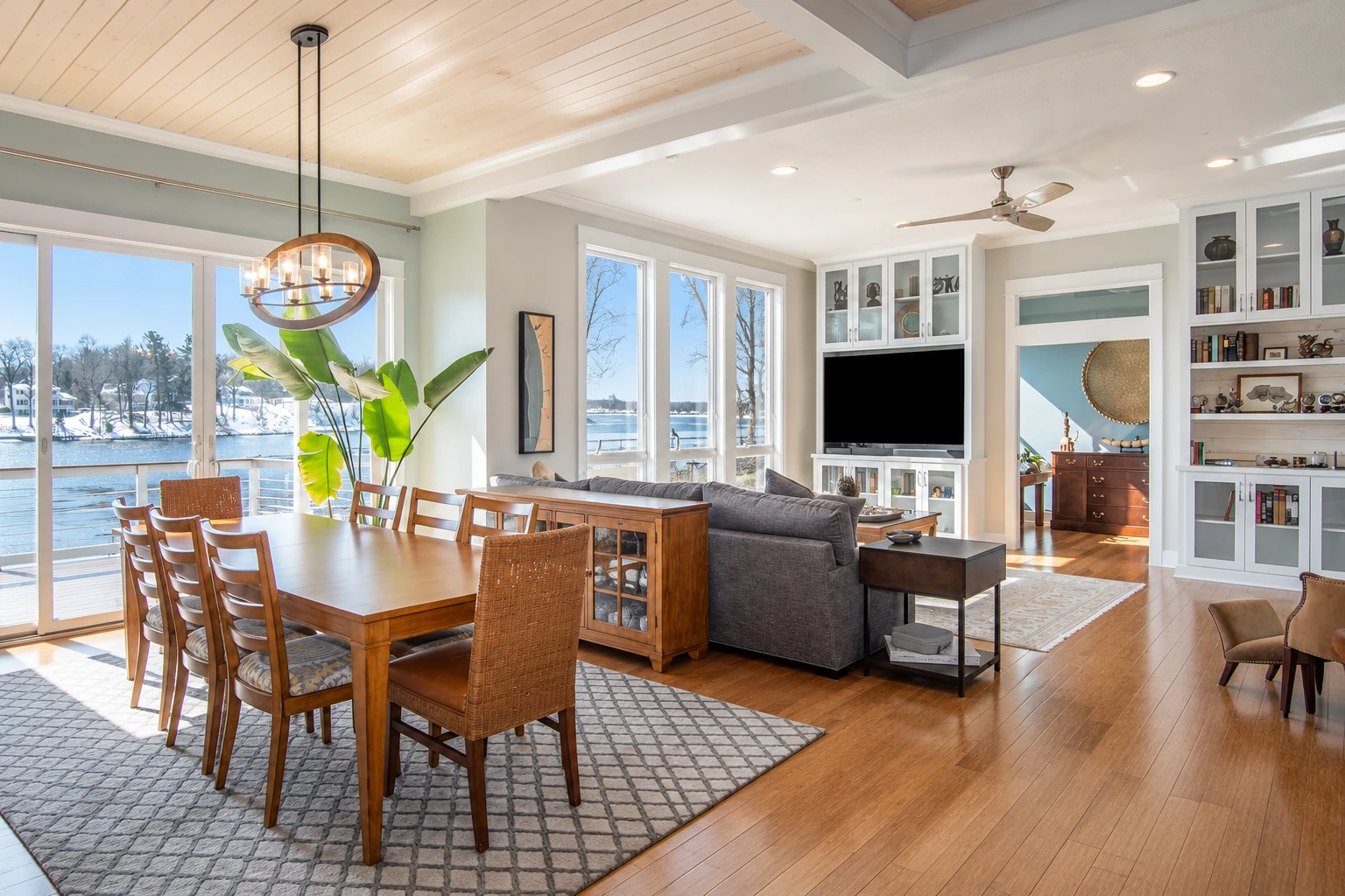Open living and dining room with large windows overlooking a lake, wooden floors and a dining table.