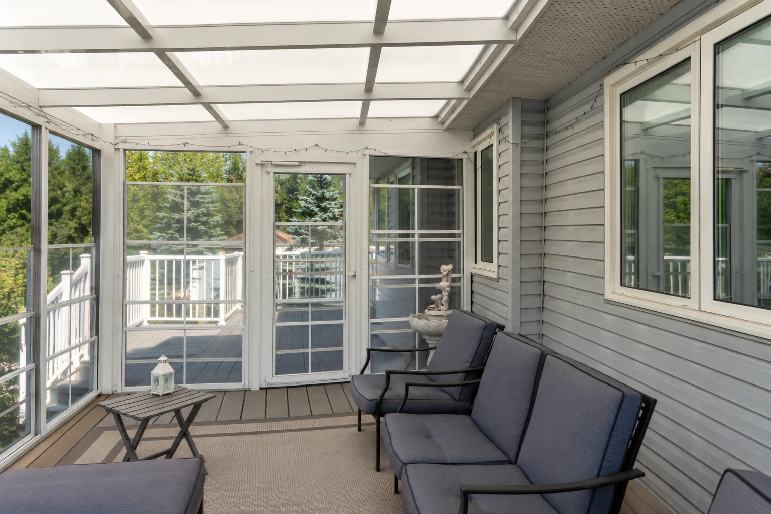 An enclosed patio with a translucent roof, white-framed windows and doors, gray siding, and cushioned chairs.
