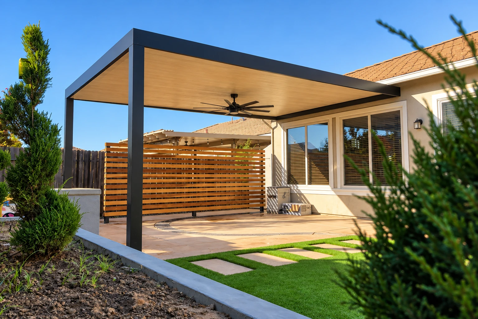 A modern patio cover with a flat wood-paneled roof, dark metal frame, and ceiling fan over a paved patio.