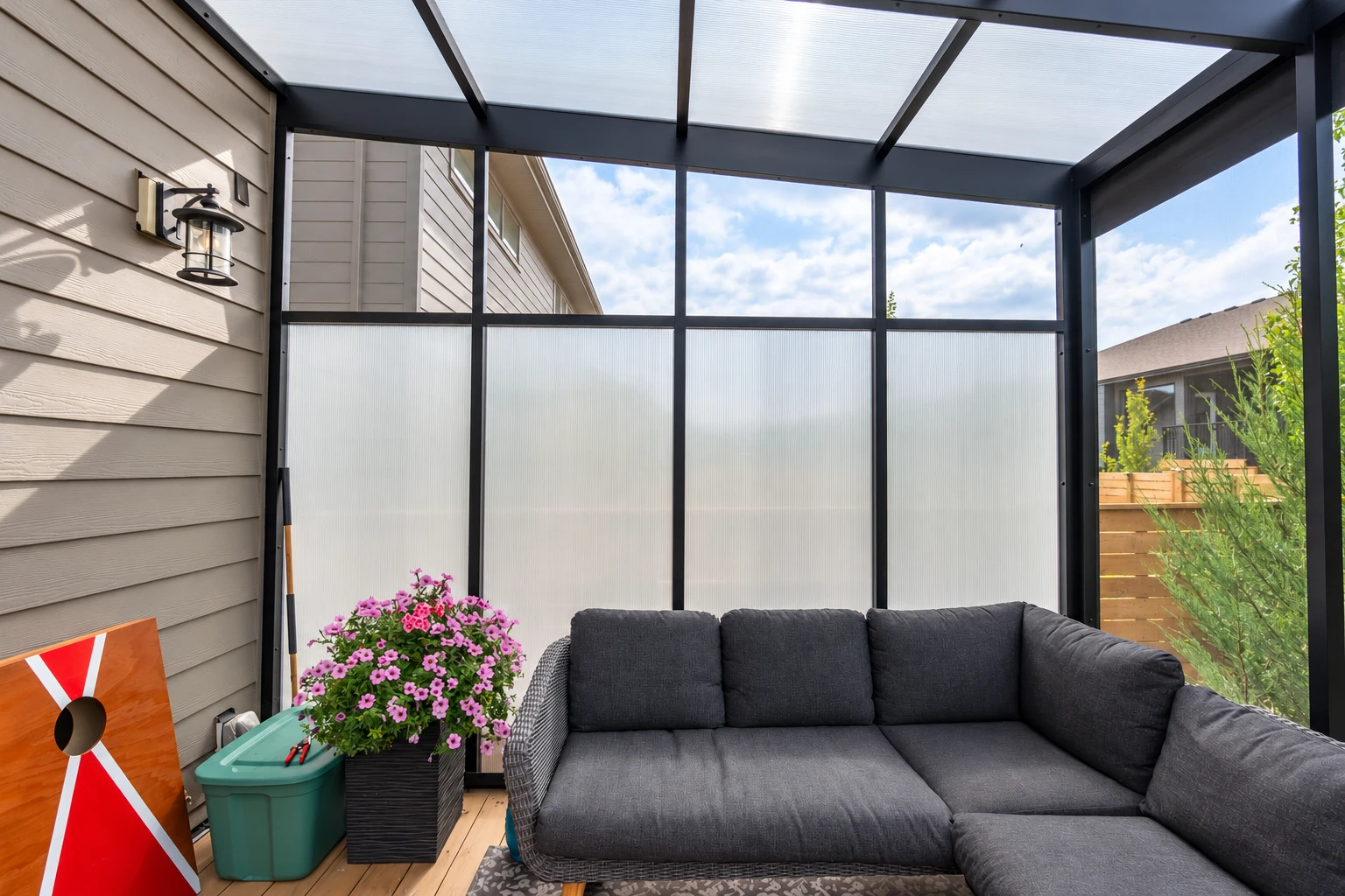 Enclosed patio with a translucent roof, dark framing, and a gray sectional beside flowering plants.