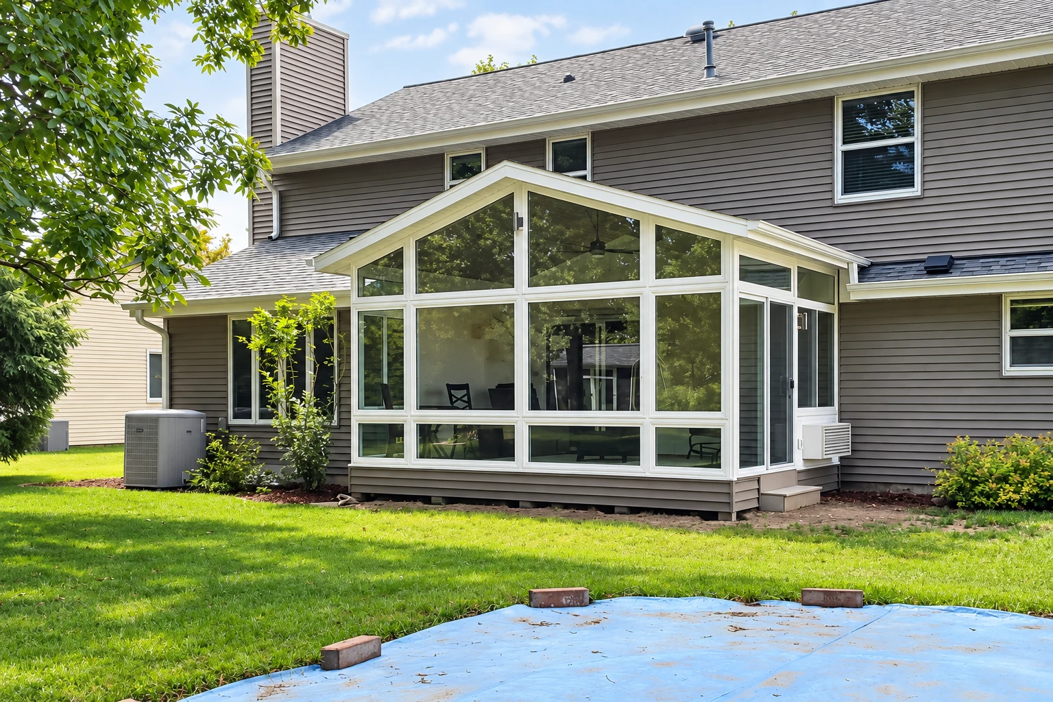 A gabled, white-framed glass sunroom projects from a gray-sided house, with a bare patio slab and lawn in front.