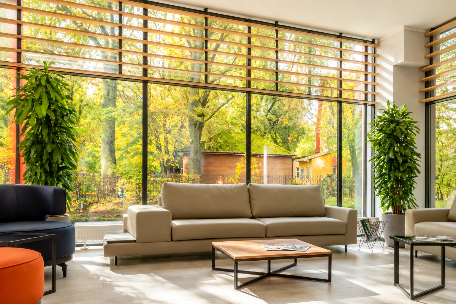 Sunroom addition with floor-to-ceiling windows, horizontal wood shades, and a beige sofa overlooking autumn trees.
