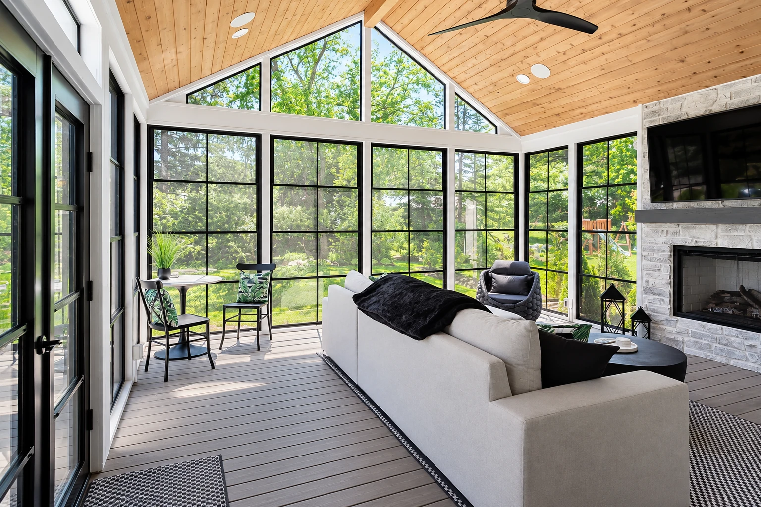 Bright sunroom interior with tall black-framed windows, a wood-paneled vaulted ceiling and a stone fireplace.