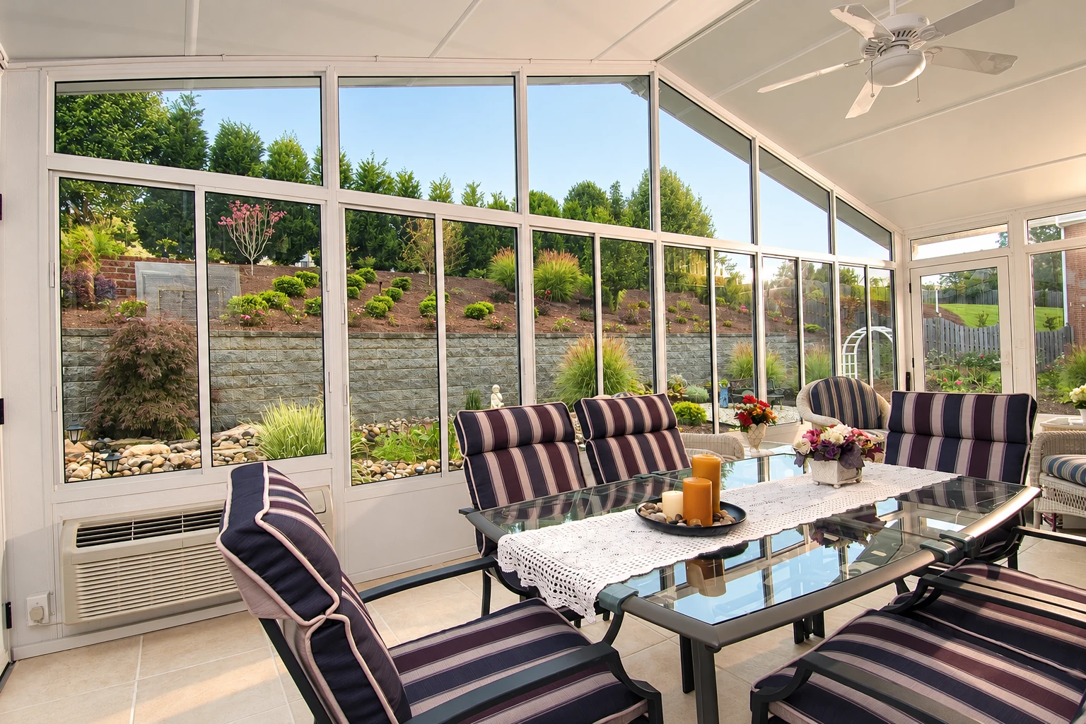 A bright sunroom with white-framed windows, striped lounge chairs, and a glass dining table faces a landscaped garden.