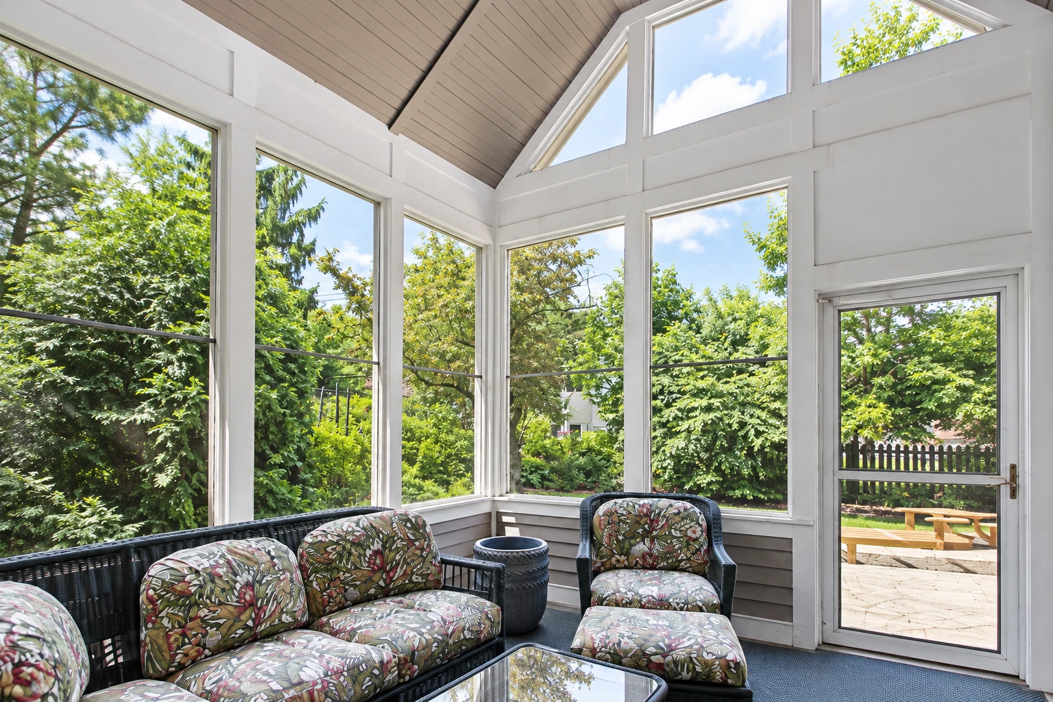 Three-season sunroom with tall windows, floral patio furniture, and a view of a leafy backyard.