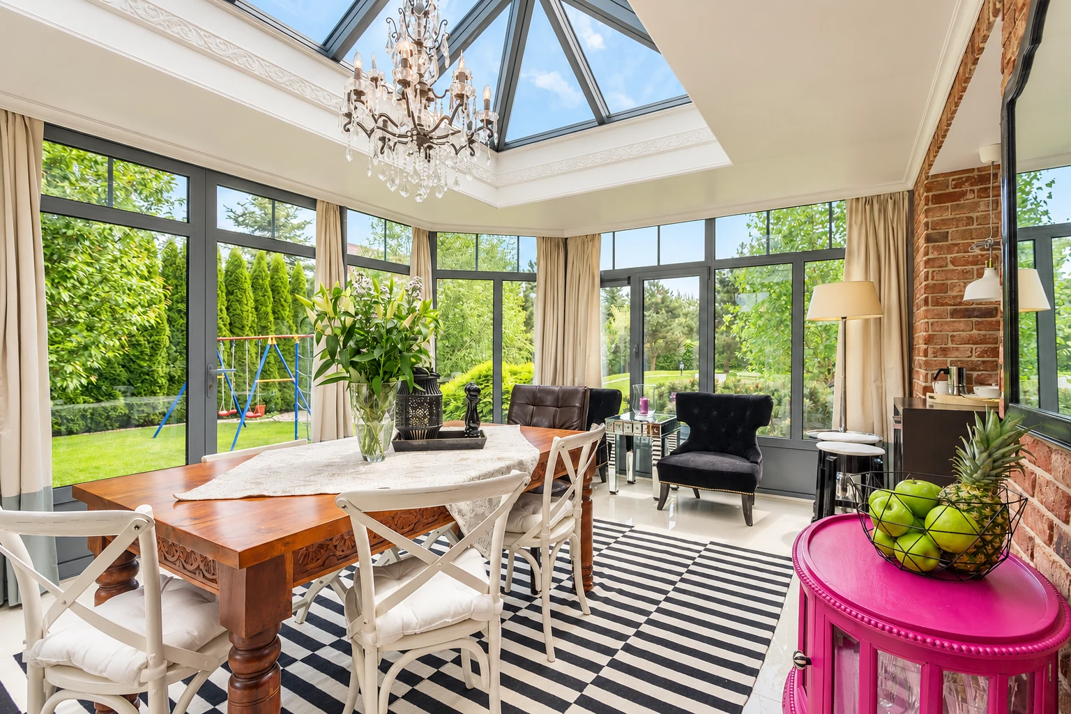A bright glass sunroom with a peaked skylight features a dining table, chairs, black-and-white rug and views of a garden.